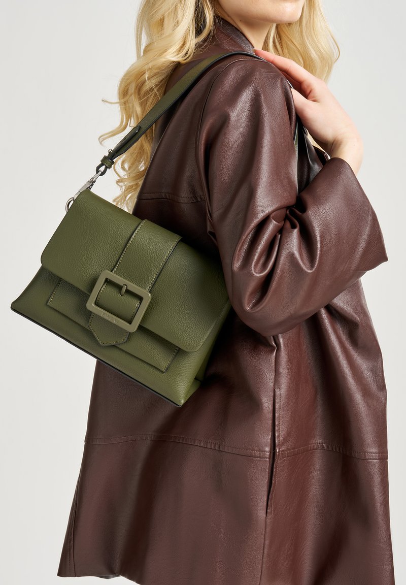 Borsa a tracolla in pelle verde oliva con una forma strutturata, caratterizzata da un pattina frontale e un dettaglio con fibbia rettangolare, indossata su una giacca marrone scuro.