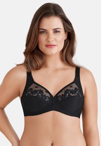 Il reggiseno nero presenta una sovrapposizione di pizzo floreale su tessuto trasparente nella parte superiore, con una sezione inferiore liscia e spalline regolabili.
