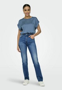 Top in satin blu con maniche corte, abbinato a jeans in denim blu a vita alta. La modella indossa sneakers bianche e semplici orecchini a cerchio in argento.