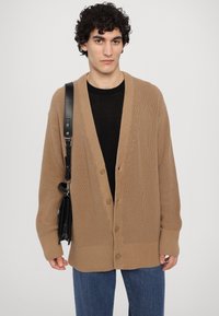 Beige strikket cardigan med V-hals, ribbestrikket tekstur og fem knapper. Brukt over en svart skjorte, kombinert med mørke denimjeans og en svart veske.