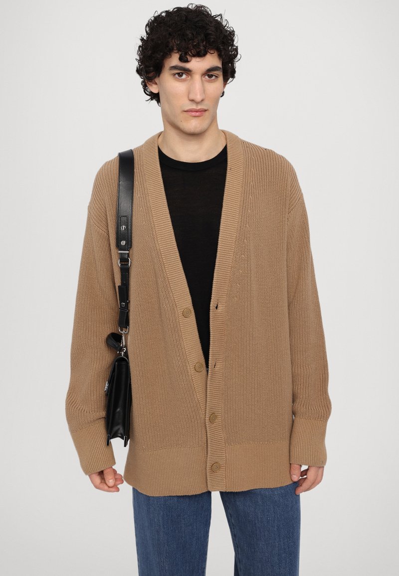 Beige gestrickte Strickjacke mit V-Ausschnitt, geripptem Muster und fünf Knöpfen. Getragen über einem schwarzen Shirt, kombiniert mit dunklen Jeans und einer schwarzen Tasche.