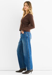 Ruskea V-puolikaulusneule, jossa on yksi nappi, yhdistettynä korkeavyötäröisiin sinisiin leveälahkeisiin farkkuihin ja teräväkärkisiin ruskeisiin kuviollisiin korkkareihin.