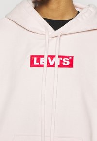 Světle růžová mikina vyrobená z měkké látky, s červeným obdélníkovým logem, na kterém je bíle napsáno "LEVI'S", a šňůrkou pro úpravu.