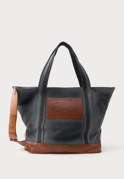 Bolso tote de lienzo en gris oscuro con detalles de cuero marrón. Presenta asas dobles y un parche frontal con texto y diseños en relieve.