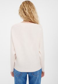 Vaaleanbeige ribbineulepaita, jossa on pitkät hihat, pyöreä kaula-aukko ja rento istuvuus, yhdistetty sinisiin farkkuihin.