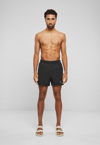 Shorts de bain noirs avec une taille élastique, des poches latérales et un logo discret. Portés avec des sandales à glisser de couleur claire sur pieds nus.
