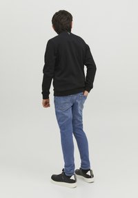 Sudadera negra con cuello redondo y puños de canalé, combinada con jeans de color azul claro que tienen un bolsillo trasero. Zapatillas negras con suelas blancas.