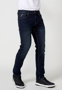 Jeans in denim blu scuro con vestibilità slim, con dettagli di cucitura, cinque tasche e una tasca con zip sul lato destro.