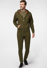 Zielona oliwkowa bluza z kapturem na zamek z czerwonymi akcentami, w połączeniu z dopasowanymi dresami. Zawiera kieszenie z przodu i czarne sneakersy. Gładka faktura materiału.