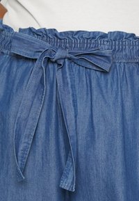 Short en denim bleu avec une taille élastique froncée, doté d'une fermeture à nouer en tissu. La texture est lisse avec une légère brillance.