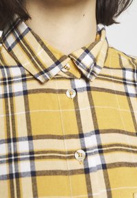 Camicia di flanella gialla a quadri con colletto classico, motivi bianchi e neri, bottoni dorati e una texture morbida.
