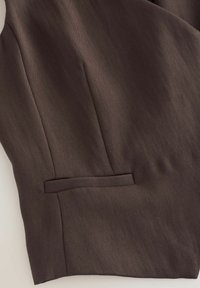 Detail hnědé střižené látky s horizontálním náprsním kapsou a viditelným šitím na hladkém texturovaném materiálu.