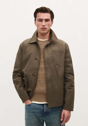 Olive green jacket with a collared design, button closure, and chest pocket, paired with a beige knitted sweater and blue jeans.