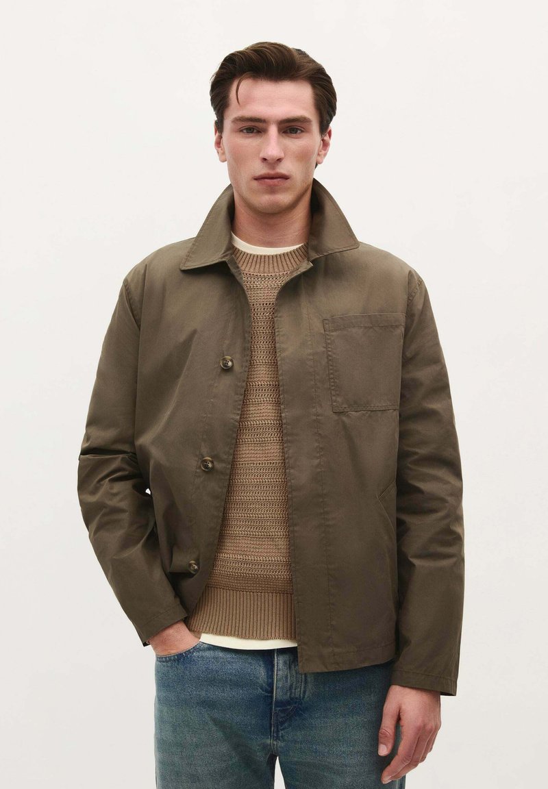 Olijfgroene jas met een kraagontwerp, knoopsluiting en borstzak, gecombineerd met een beige gebreide trui en blauwe jeans.