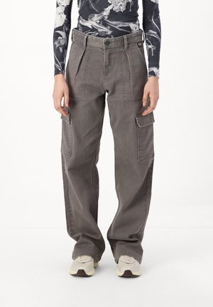 Osoba w luźnych szarych spodniach typu cargo z bocznymi kieszeniami i białych trampkach, stojąca na tle jednolitego białego tła.