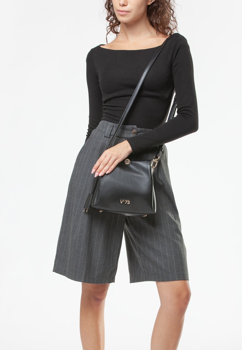 Bolso de cuero negro con correa ajustable, forma rectangular, logo dorado "V-73," combinado con pantalones cortos de talle alto a rayas grises y una blusa negra.