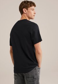 Jaunas vyras su garbanotais rudais plaukais, vilkintis paprastą juodą marškinėlį ir išblukusius pilkus džinsus, stovi su rankomis kišenėse, pasukęs į šoną.