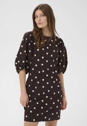 Ženska v črni obleki z belimi pikami in napihnjenimi rokavi do komolcev, stoji ob enobarvnem ozadju.