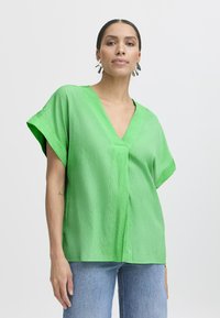 Blusa verde a maniche corte con scollo a V in tessuto testurizzato, vestibilità morbida e cucitura centrale. Indossata con jeans azzurri chiari.