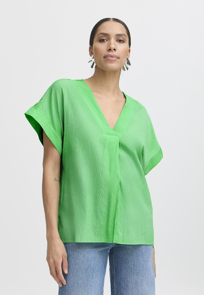 Blusa verde a maniche corte con scollo a V in tessuto testurizzato, vestibilità morbida e cucitura centrale. Indossata con jeans azzurri chiari.