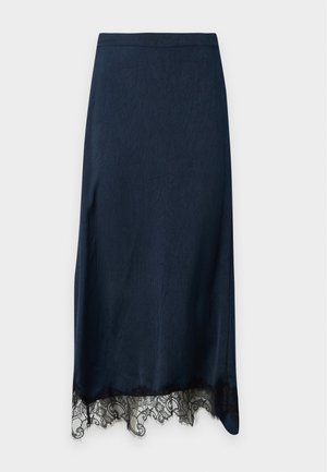 Femme portant une chemise à rayures bleues boutonnée, une jupe midi foncée avec un ourlet en dentelle et des chaussures plates bordeaux, debout, une main sur la hanche.