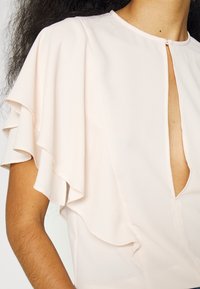 Blouse beige clair avec manches volantées superposées et encolure fendue à l’avant portée par une personne aux cheveux bouclés.