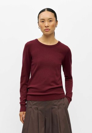 Kvinde med glat sort hår i en lav hestehale, iført guldringeøreringe, en burgunderrød langærmet sweater og en brun plisseret nederdel.