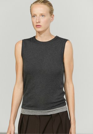 Donna con capelli biondi raccolti indietro che indossa una canotta grigio scuro sopra una maglietta grigio chiaro e pantaloni marrone scuro su uno sfondo semplice.