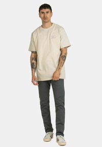 Beige Baumwoll-T-Shirt mit kurzen Ärmeln und einem auf der linken Brust gedruckten "REELL"-Logo, kombiniert mit dunkelgrauen Hosen und weißen Sneakers.