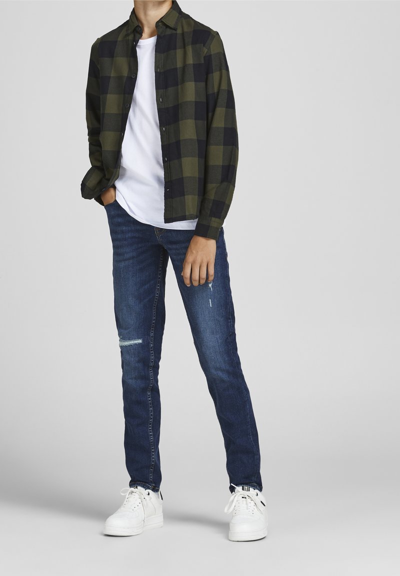 Chemise à carreaux verts et noirs sur un t-shirt blanc, associée à un jean déchiré bleu foncé et des baskets blanches, avec des lacets et un tissu texturé.