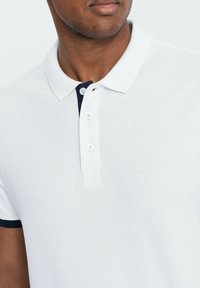 Polo shirt bianco a maniche corte realizzato in tessuto testurizzato, con regolazione a tre bottoni e dettagli blu navy sul colletto e sulle maniche.