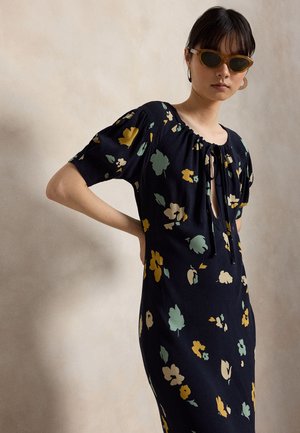 Kvinde iført sort blomsterkjole med pufærmer og bindedetalje foran, accessoriseret med brune cat-eye solbriller og store hoops-øreringe.