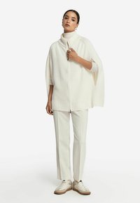 Krämfärgad stickad poncho med hög krage och dragkedja framtill, med breda ärmar och ribbad nederkant. Parat med beiga byxor och sneakers.