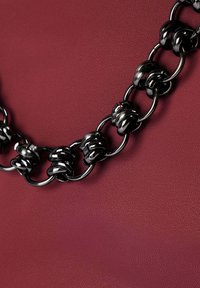 Un lanț de metal negru cu verigi răsucite, expus pe un fundal texturat bordeaux. Finisaj neted și lucios cu accesorii circulare.