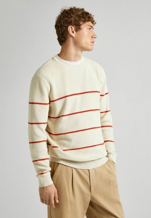Suéter de punto acanalado crema con rayas horizontales rojas, cuello redondo, mangas largas y un corte relajado. Combinado con pantalones beige de pinzas.