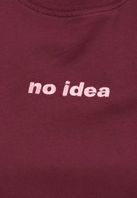 Μπορντό ύφασμα με κεντητό κείμενο σε ανοιχτό ροζ χρώμα, «no idea» με μικρά γράμματα, στο κέντρο ενός ενδύματος.