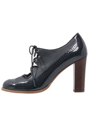 Zapato de charol negro con cierre de cordones, detalles de brogue, punta almendrada y tacón medio de madera. Suela texturizada para mayor agarre.