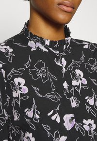 Blusa nera con colletto a volant, caratterizzata da un motivo floreale in lavanda e bianco, e bottoni neri lungo la parte anteriore.