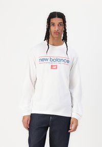 Hvit langermet T-skjorte med "new balance" logo i rødt og blått. Har rund hals og avslappet passform. Kombinert med mørk denimjeans.