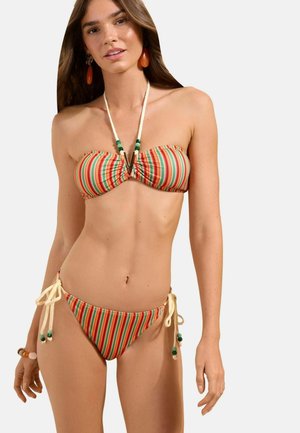 Kobieta nosząca wielokolorowy, paskowany bikini typu bandeau z wiązaniami z liny i dekoracyjnymi zielonymi koralikami, uzupełniona dużymi kolczykami i bransoletkami.
