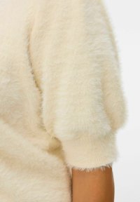Puszysty sweter w kremowym kolorze z krótkimi, puszystymi rękawami i miękką teksturą. Tkanina wydaje się lekka i ma luźny krój.