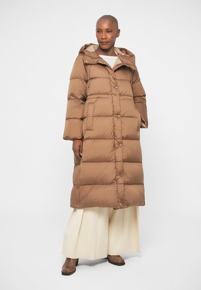 Brun pufferjakke med hætte, der har en knappet front, store lommer og et quiltet design. Bærer den over cremefarvede bukser med bredde.