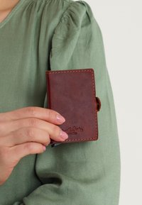 Bruine leren portemonnee met zichtbare stiknaden, vastgehouden in een hand. Kenmerkt zich door een reliëfmerk aan de voorkant en afgeronde hoeken. Compact ontwerp.