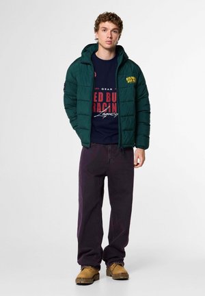 Junger Mann trägt eine grüne Steppjacke, ein dunkelblaues Red Bull Racing Sweatshirt, dunkle Hosen und braune Arbeitsstiefel und steht vor einem schlichten Hintergrund.