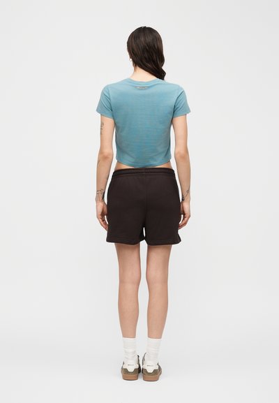 Γυναίκα που φαίνεται από πίσω φοράει μπλε κοντομάνικο μπλουζάκι, μαύρο σορτς, λευκές κάλτσες και αθλητικά παπούτσια, με φόντο έναν απλό φωτεινό τοίχο.