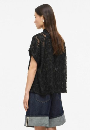 Sieviete ar gariem brūniem matiem valkā melnu teksturētu caurspīdīgu topu un uzvērstas tumšas džinsa šortus, raugoties pa kreisi uz neitrālu fonu.