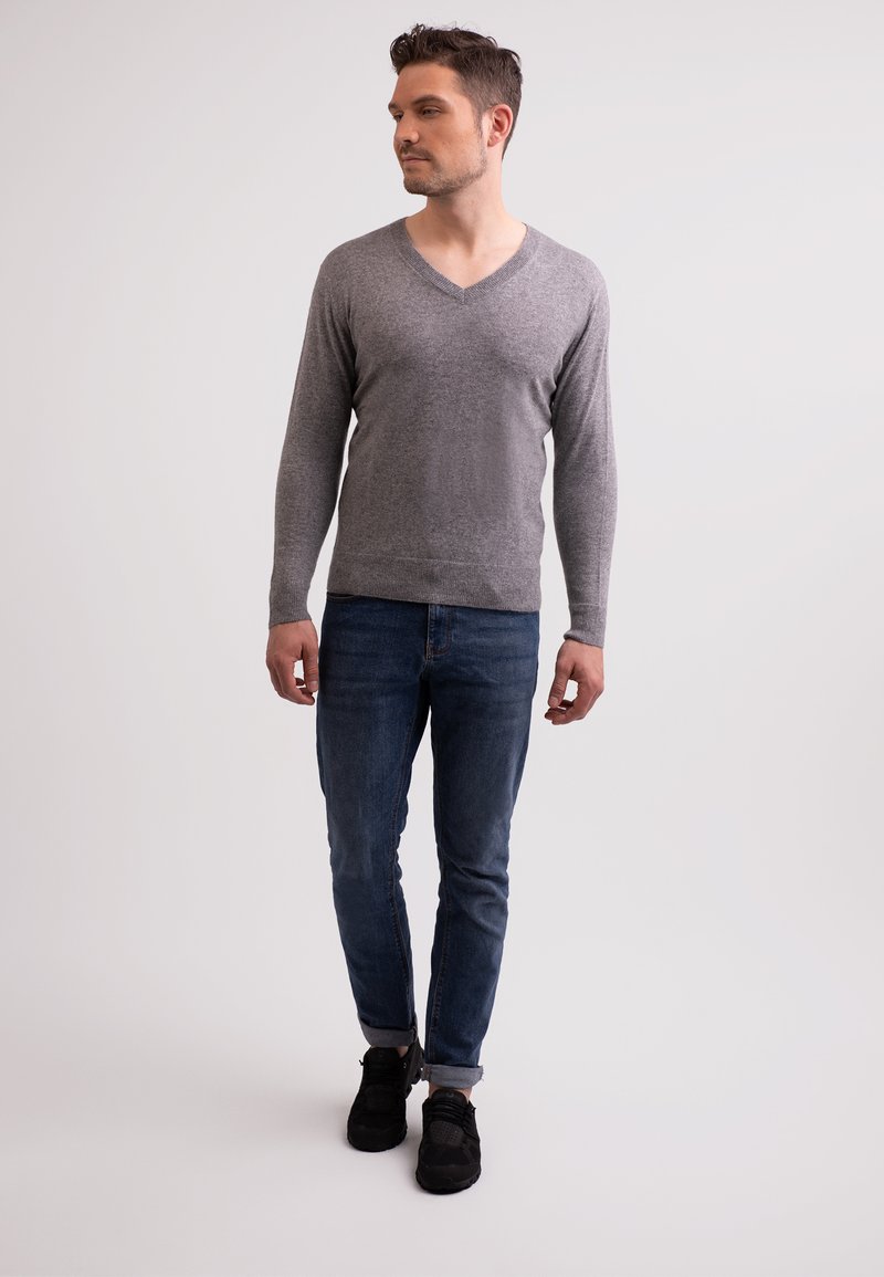 Szara, długosi sleeve sweter z dekoltem w serek, miękka tekstura, w zestawieniu z niebieskimi dżinsami oraz czarnymi sneakersami z widocznymi sznurowadłami i fakturowaną powierzchnią.