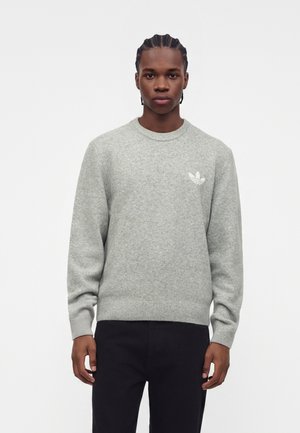 Joven con el pelo trenzado que lleva un suéter gris claro con cuello redondo con un pequeño logo blanco en el pecho izquierdo y pantalones negros.