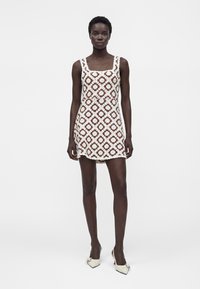 Femme debout portant une mini-robe sans manches en crochet marron et blanc avec des talons blancs à bout pointu sur un fond uni.