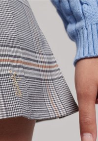 Falda a cuadros en gris, blanco y naranja, con un diseño plisado y un logotipo bordado en dorado cerca del dobladillo. Manga de suéter de punto azul visible.